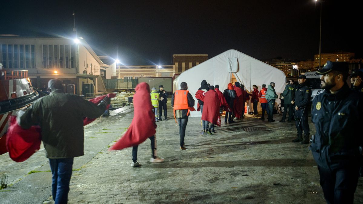 Gerettete Migrantinnen und Migranten warten vor einem Zelt des Roten Kreuzes im Hafen von Málaga auf ihre medizinische Versorgung. Menschen mit roten Decken stehen unter künstlicher Beleuchtung vor einem weißen Zelt.